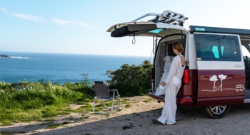 Woman leaning on red and white campervan staring at the sea in front of her.