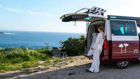 Woman leaning on red and white campervan staring at the sea in front of her.