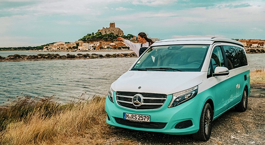 Green Mercedes Vito camper standing at a beach with city in the back
