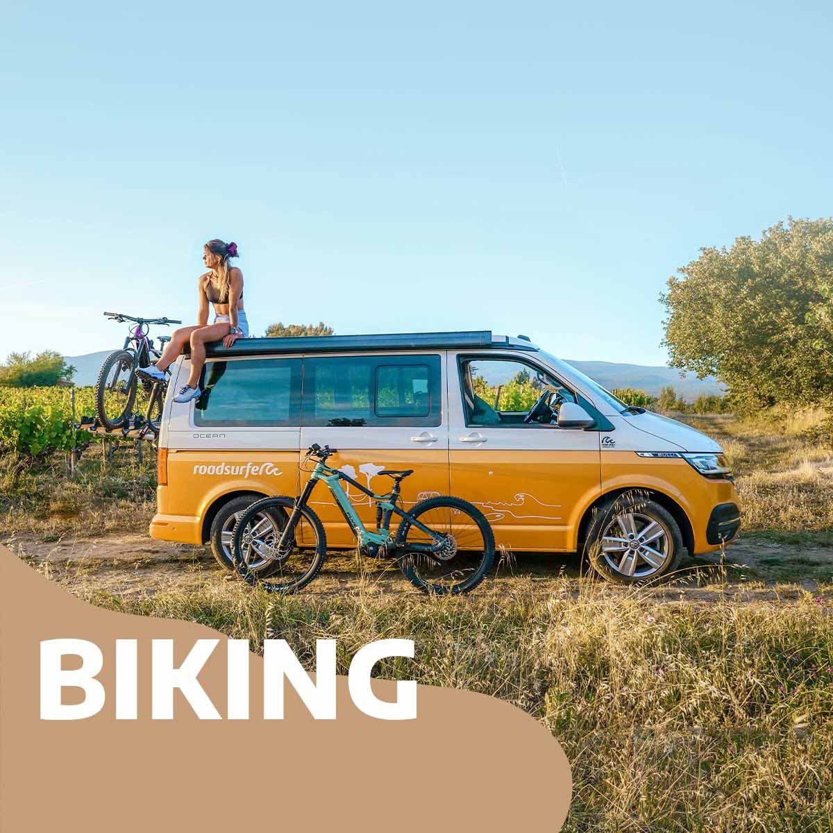 Woman sits on top of a VW California campervan with a bike leaning against the vehicle