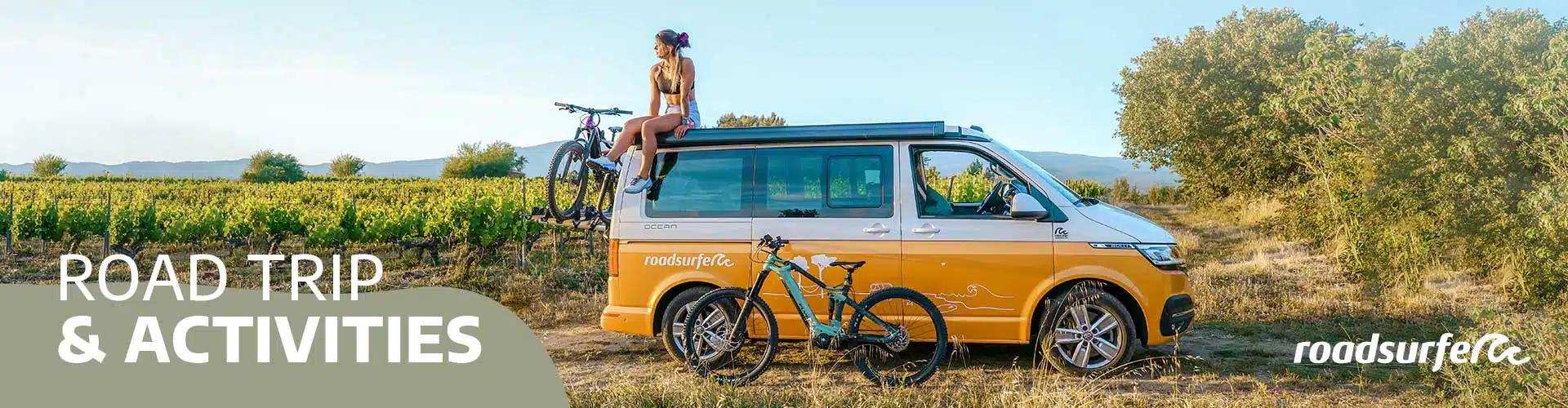 girl sitting on top of a campervan on a field with bikes next to it