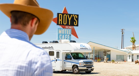 Man with cowboy hat walking towards an RV parked in front of a motel.