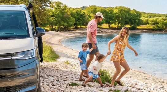 Family of four holding hands and walking from their campervan towards the lake.
