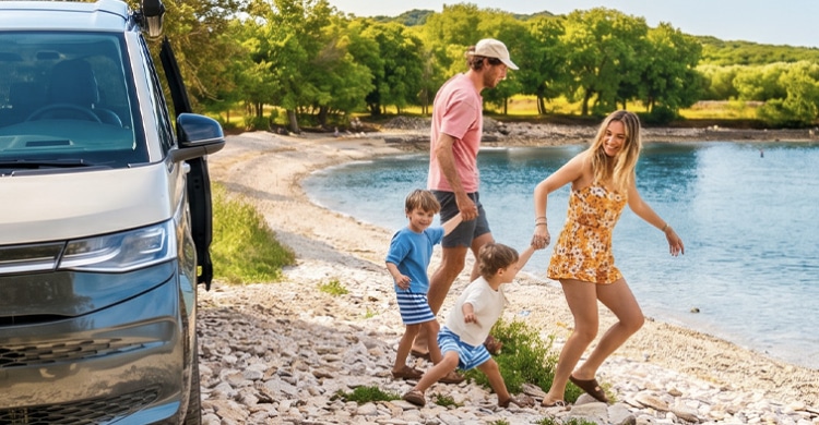 Family of four holding hands and walking from their campervan towards the lake.