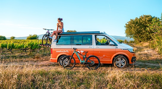 girl sitting on top of an orange campervan equipped with bikes and parked in the vineyards