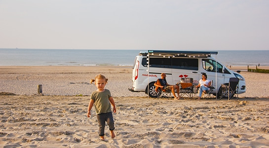 Ford Nugget Camper parked on the sand of a beach and a little girl playing in front of it