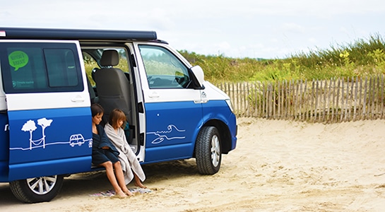 Kids sitting in a blue campervan from roadsurfer at a beach