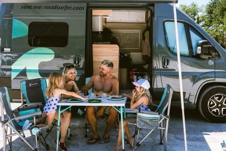 Family enjoying a meal outdoors at a table beside their camper van on a sunny camping day.