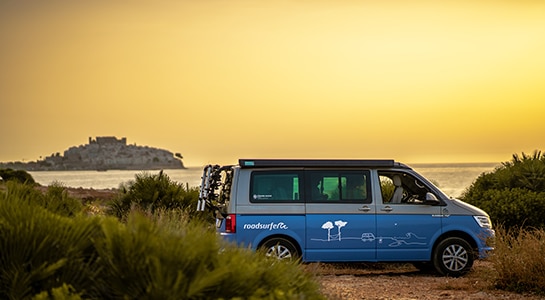 Blue Volkswagen California camper standing at the seaside with sundown in the background