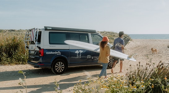 A group of friends parks their VW camper by the beach and heads toward the sea with their surfboards.