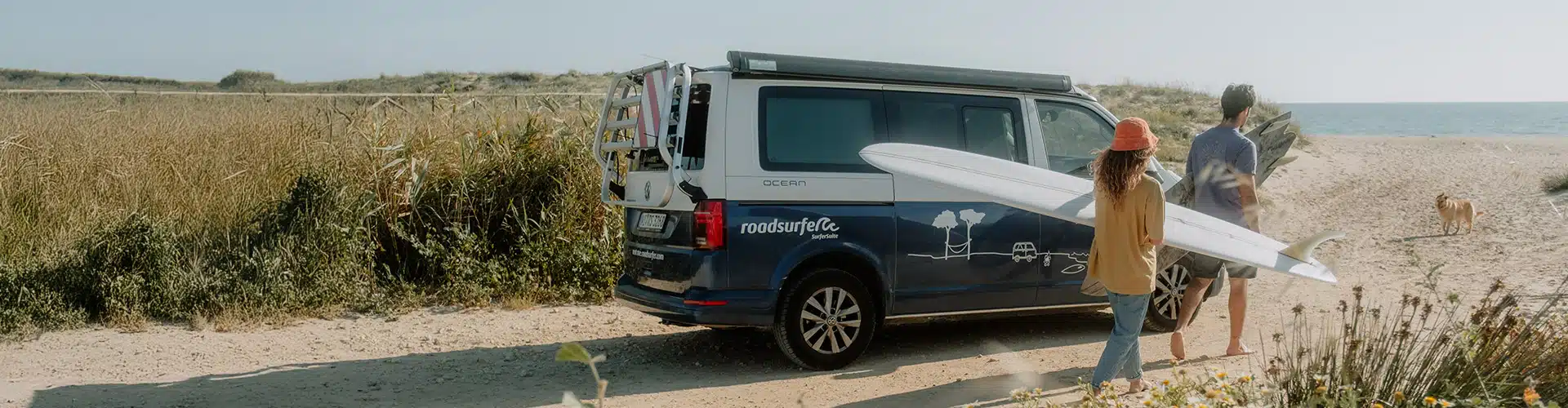 A group of friends parks their VW camper by the beach and heads toward the sea with their surfboards.
