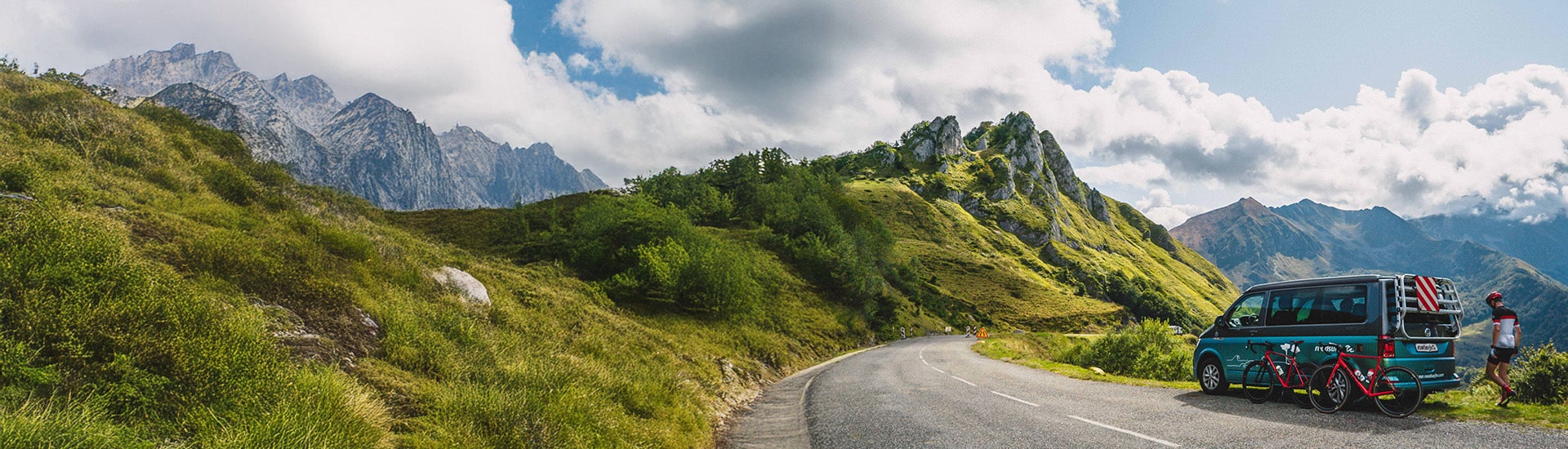 Biker walking behind hier parked campervan at scenic mountain road with two bikes leaning against the vehicle 