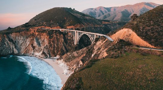 View of Bixby Creek Bridge arching over rugged cliffs on California’s Big Sur coast, with waves rolling onto a secluded beach below.