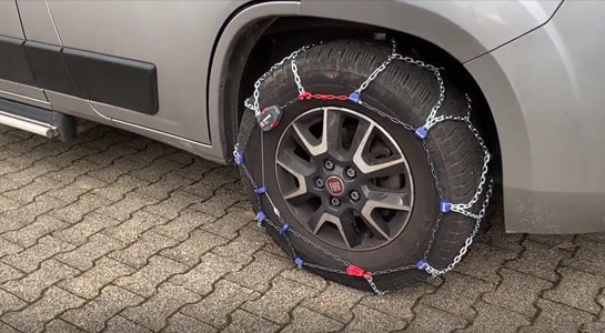 Snow chains around the right front wheel of a grey campervan.