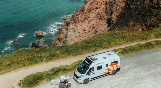 Boxvan parked at the edge of a cliff with people sitting next to it around a camping table.