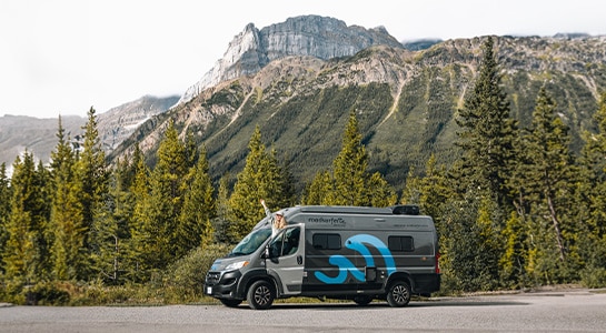 Woman leaning out of a boxvan parked at the side of the road with the mountains visible in the background.