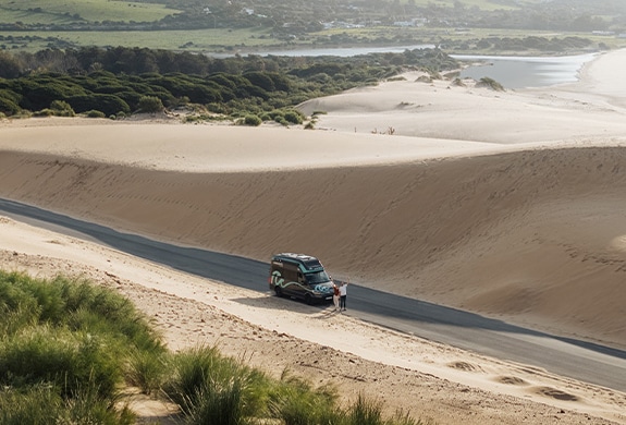 Boxvan parked on the route in between dunes with two people standing and waving at the camera capturing the picture from above.