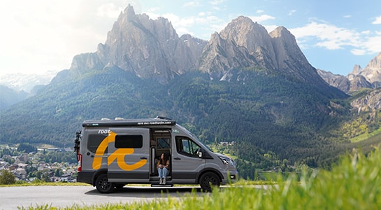 Boxvan parked with view over the Dolomites in the background, while a person is sitting in the doorway of the van.