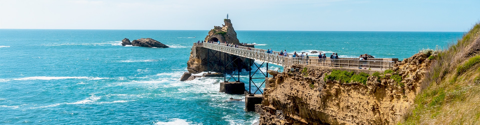 Bridge leading towards cliff in the sea with people walking over it.