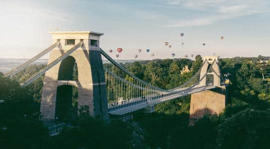 Hot air balloons floating above the Clifton Suspension Bridge in Bristol, United Kingdom, during sunrise.