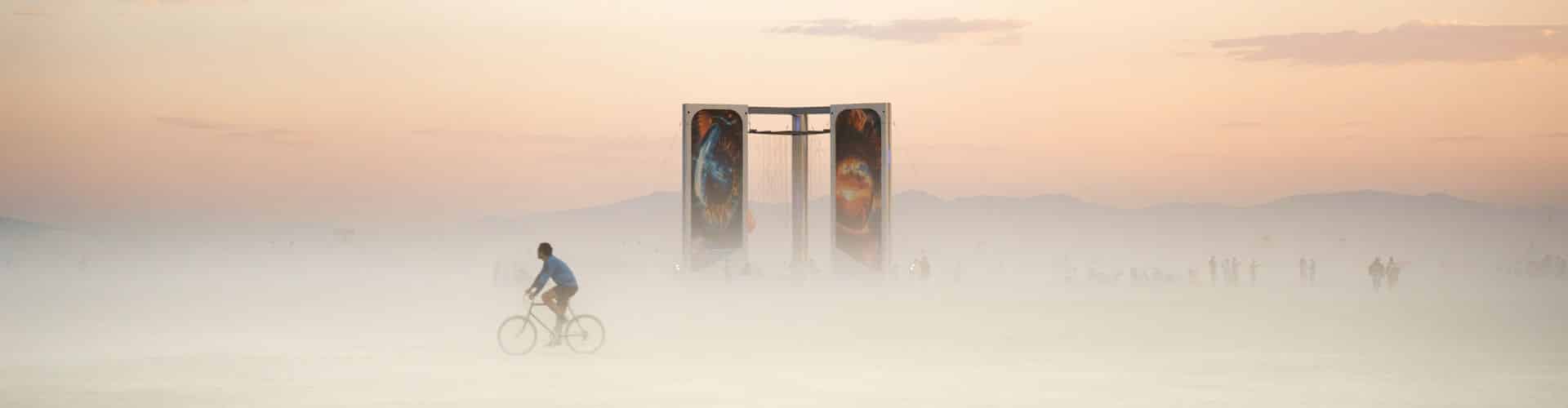 Cyclist riding through low dust at sunrise past a tall art installation with painted panels at Burning Man