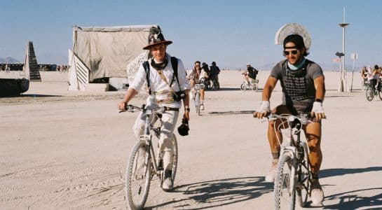 Two people in costume riding bikes along a dusty road at Burning Man under a clear blue sky