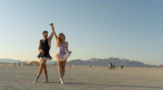 Two people in tutus dancing on the open playa at sunset with mountains on the horizon at Burning Man