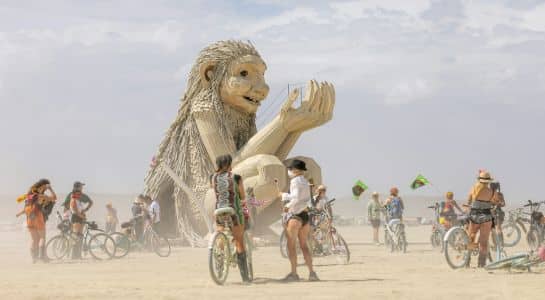 Cyclists gathered in blowing dust below a giant wooden lion sculpture at Burning Man