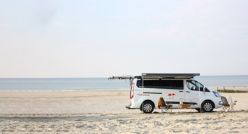Camper Cabin Deluxe camper van from roadsurfer parked on an empty beach on a sunny day, awning open, table and chairs underneath