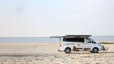 Camper Cabin Deluxe camper van from roadsurfer parked on an empty beach on a sunny day, awning open, table and chairs underneath