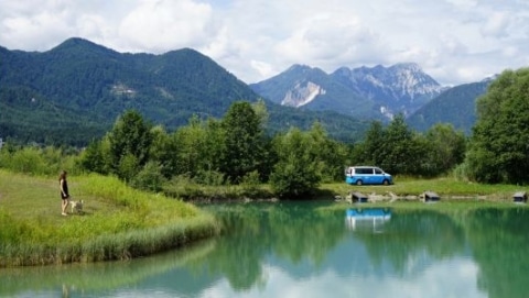 Blue camper van parked near a calm lake with mountains and trees in the background