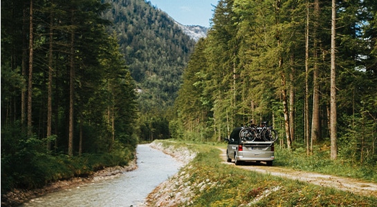 Grey campervan driving through the forest next to a river.