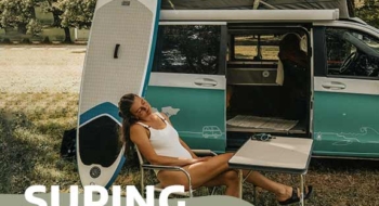 Frau in einem Campingstuhl, neben Campervan mit SUP-Board am Van angelehnt