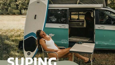 Frau in einem Campingstuhl, neben Campervan mit SUP-Board am Van angelehnt