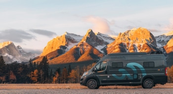 Camper van parked in front of a breathtaking mountain range bathed in warm sunlight, surrounded by evergreen trees and autumn foliage