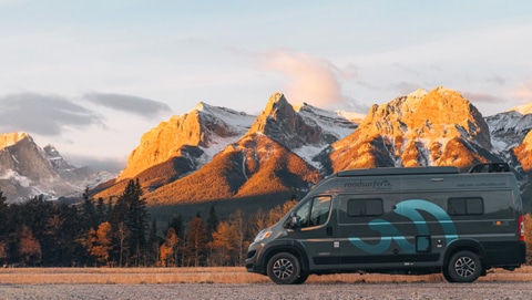 Camper van parked in front of a breathtaking mountain range bathed in warm sunlight, surrounded by evergreen trees and autumn foliage