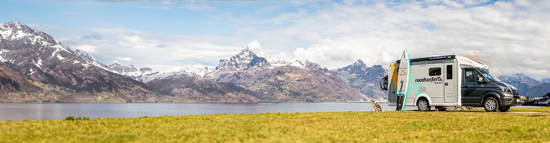 camper van parked on a grassy field near a lake, with a backdrop of majestic mountains, a partly cloudy sky, and a surfboard attached to the vehicle, evoking a sense of outdoor adventure and travel.