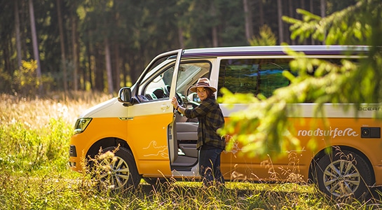 Woman standing at the door of her yellow VW camper in a forest