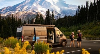 Two travelers standing near a camper van with mountains and wildflowers in the background