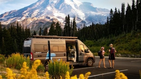 Two travelers standing near a camper van with mountains and wildflowers in the background