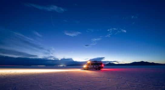 Campervan parked on white salt flats at night, headlights casting a long light trail under a deep blue sky