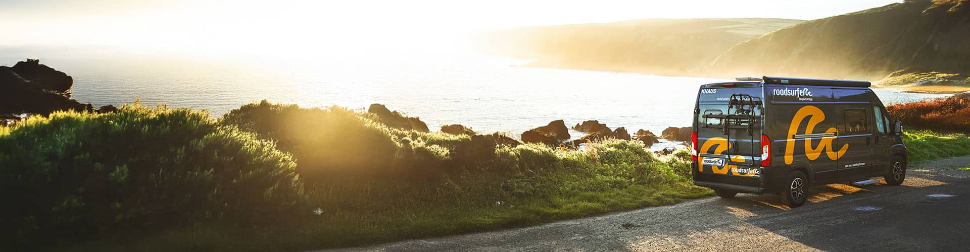 ampervan parked on a scenic coastal road with lush greenery, overlooking the ocean at sunset