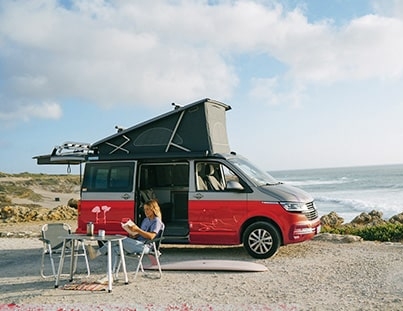 camper van with a pop-up roof parked by the ocean, where a woman is sitting at a portable table reading a book, enjoying the scenic coastal view
