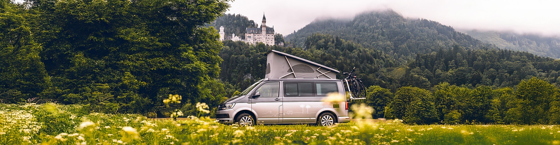 Grey campervan parked in a valley, with the castle of Neuschwanstein visible on a hill in the background.