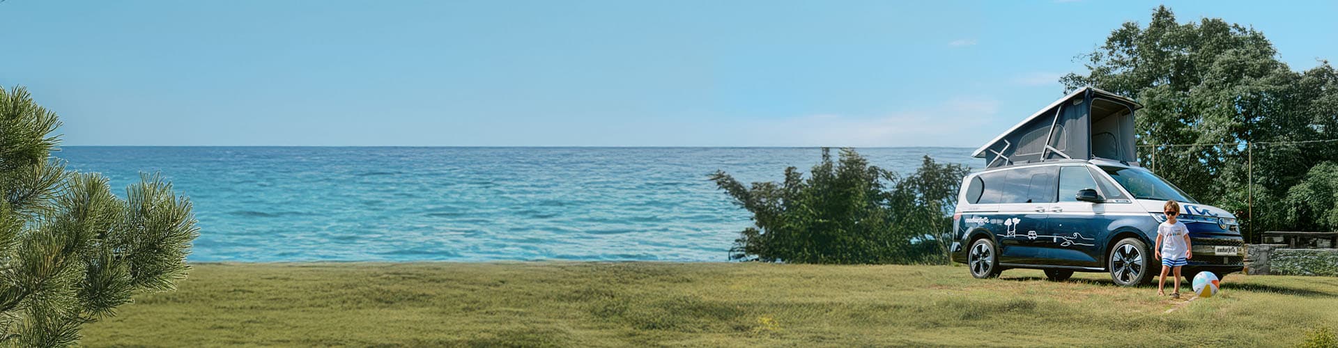 camper van with a pop-up roof is parked near the ocean on a grassy area, where a child is playing with a beach ball under a clear blue sky