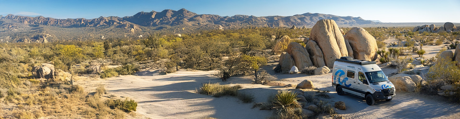 A camper van is parked on a sandy desert trail surrounded by large boulders, sparse vegetation, and distant mountain ranges under a clear blue sky