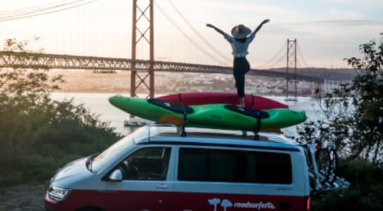a woman stands on the roof of a VW campervan with two kayaks and watches the sunset