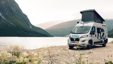 camper van with a pop-up roof parked by a scenic lake, surrounded by mountains and lush greenery, creating a tranquil outdoor setting