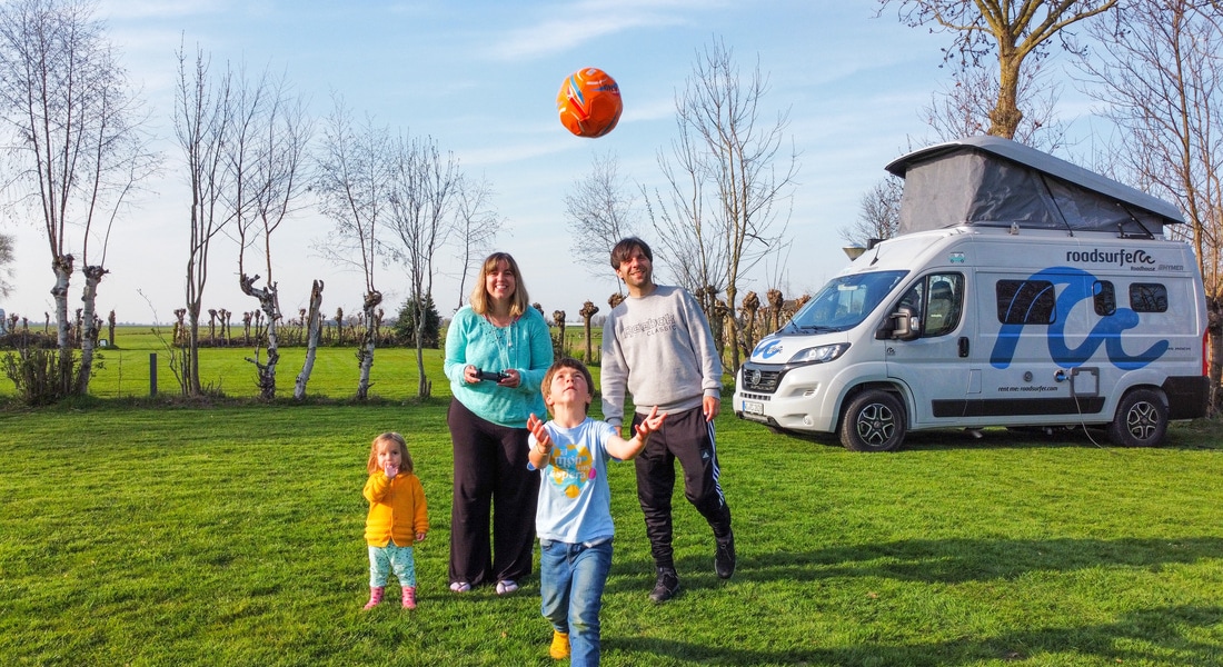 Family with 2 children playing ball in front of a campervan standing on a green field