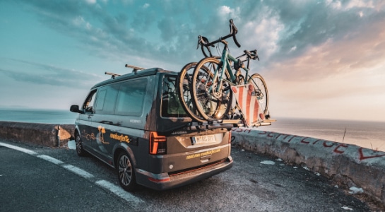 Grey camper with a bicycle rack standing on a parking lot next to a road overlooking the sea, sunset scenery